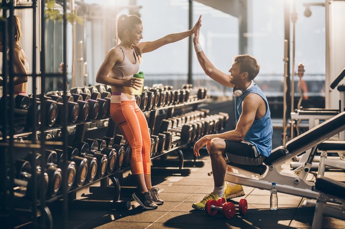 A man and woman in workout clothing high-fiving with exercise equipment in the background.