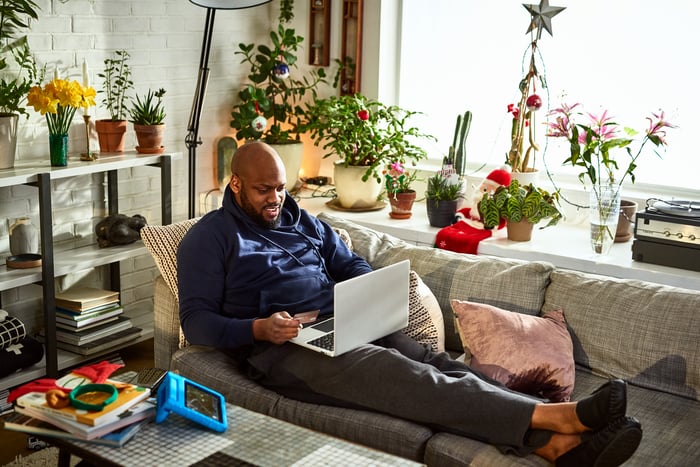 Person lounging on couch at home holds a credit card in one hand while browsing on a laptop.