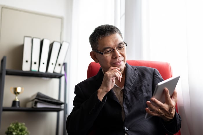 Middle-aged person sits in a red chair, looking at a tablet in one hand.