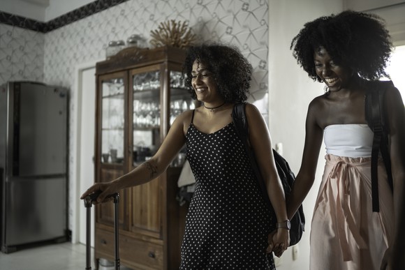 Two smiling women bring their luggage into a room.