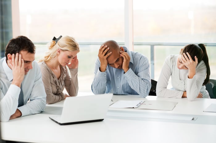 A group of four businesspeople express frustration while one talks on the phone and others look at laptops and papers.