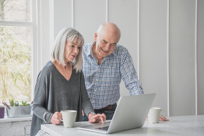 An older couple in a kitchen looking at a laptop while drinking coffee. 