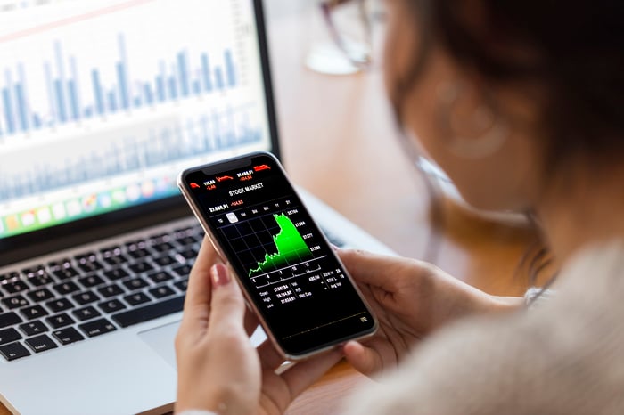 A person looking at a rapidly rising stock chart on their smartphone, with an open laptop in front of them.