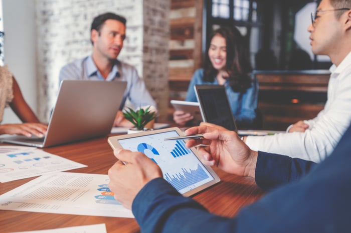 A group of people in a business meeting with laptops, tablets, and documents displaying operating metrics.