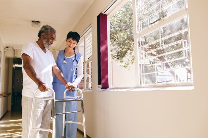 A person in medical scrubs helping another person using a walker.