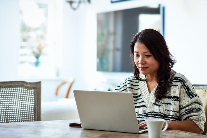 Mature person sitting at a table with a mug in front of them uses a laptop.