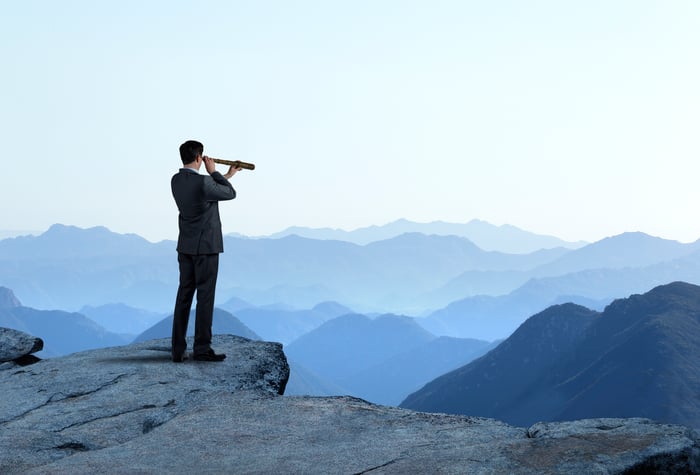 A person standing on a cliff uses a telescope to look out toward a horizon full of mountain ranges.