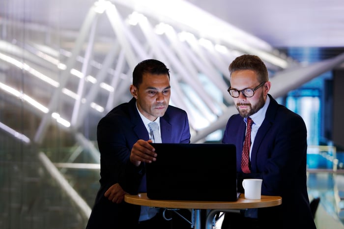 Two professionals dressed in suits look over the same laptop at a high top table.