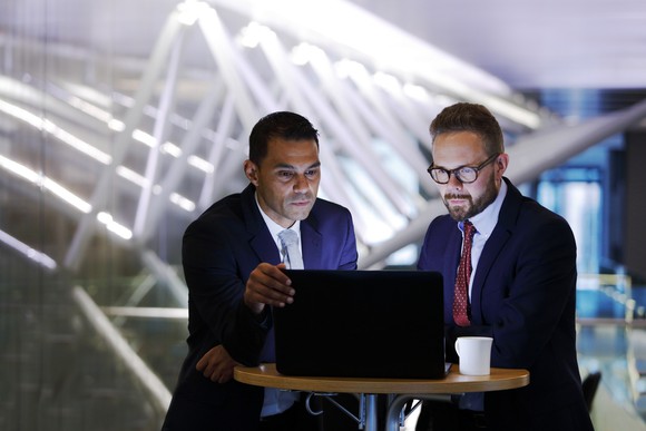 Two professionals dressed in suits look over the same laptop at a high top table.