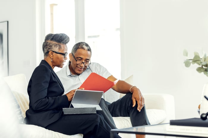 Two older people sit on a couch while reviewing documents.