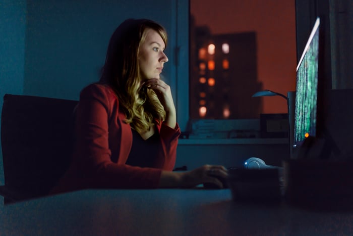 Person sitting at a desk in a dimly lit room works on code displayed on a PC.