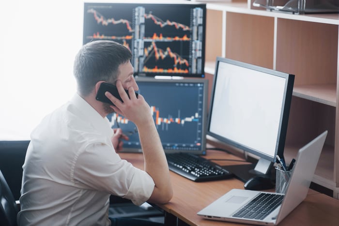 A person on phone sitting at a table with multiple computer screens displaying charts and data.