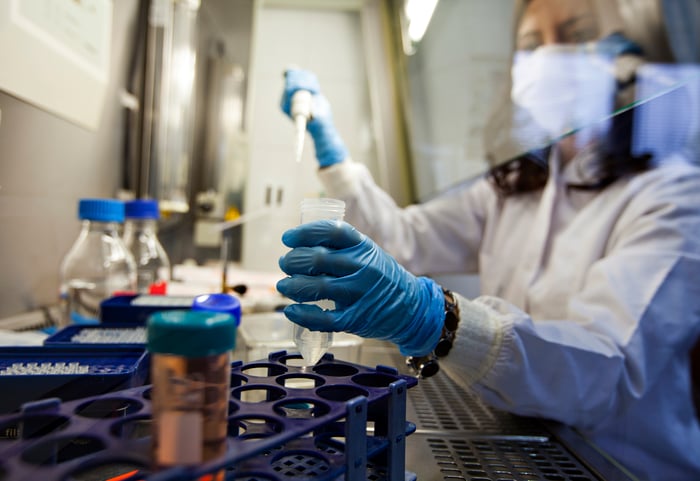 Scientist wearing a white lab coat, gloves, and mask works in a lab setting.