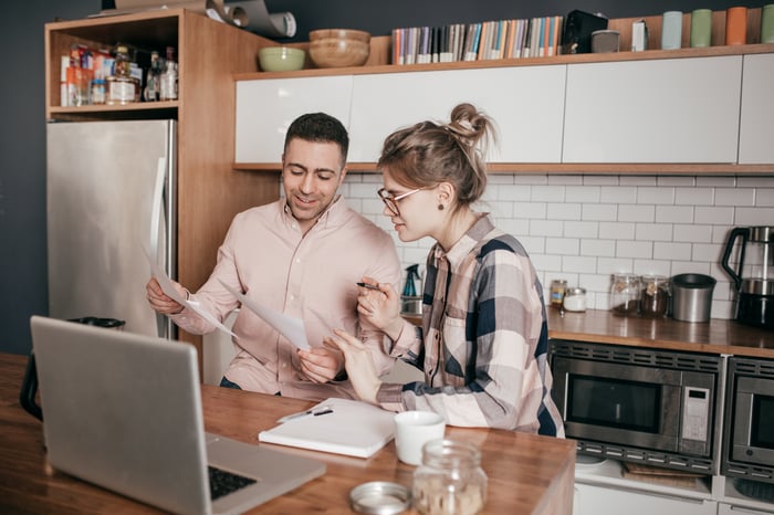 Two people review documents in front of a laptop at a kitchen counter.