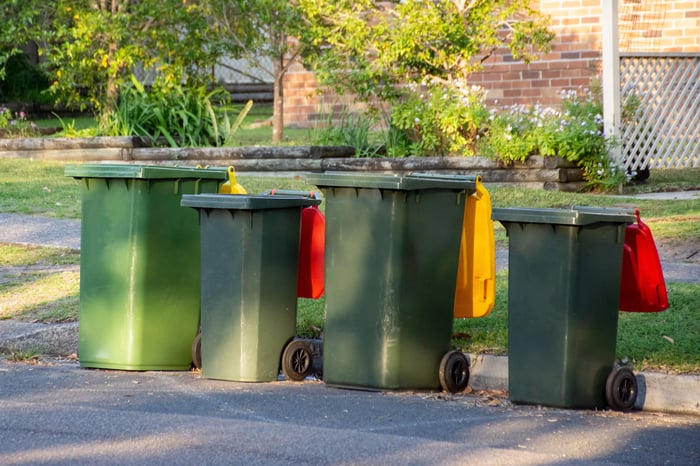 Four dark green trash cans on the side of a street in front of what appears to be a house.