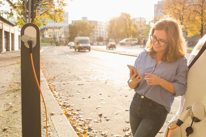 Smiling young person with a smartphone in hand is leaning against an electric car as it charges.