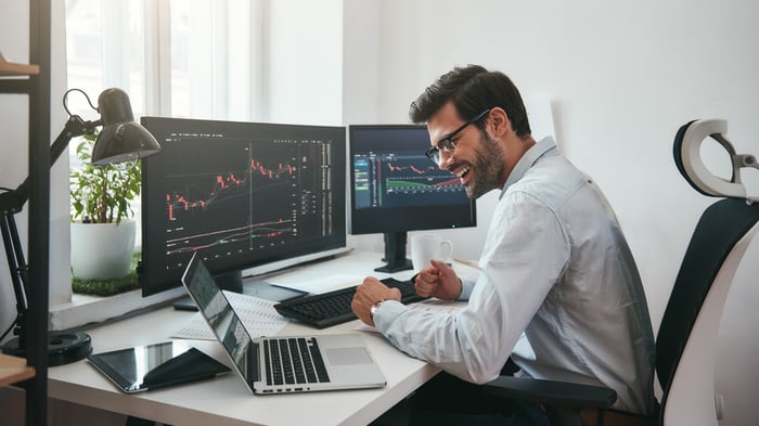 A happy investor sits at a desk and looks at financial charts on several computers.