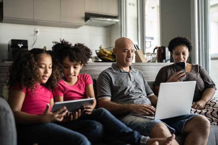 A family sits on a couch while the father works on his laptop, the mother looks at her phone, and the two children share a tablet.