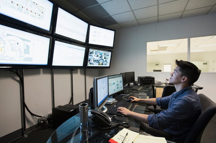 A man sits at a computer looking at a wall packed with multiple monitors.