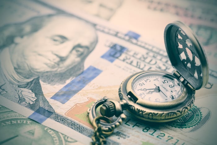 An open antique pocket watch placed atop a neatly fanned pile of one hundred dollar bills. 