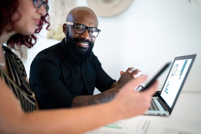 Two investors consider readouts on a laptop and a phone while sitting side by side as one smiles.