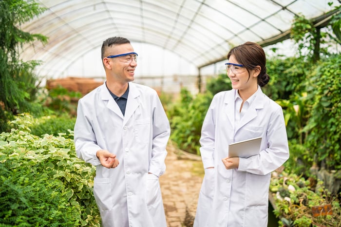 Two people in lab coats and goggles smile at each other while working inside a greenhouse.