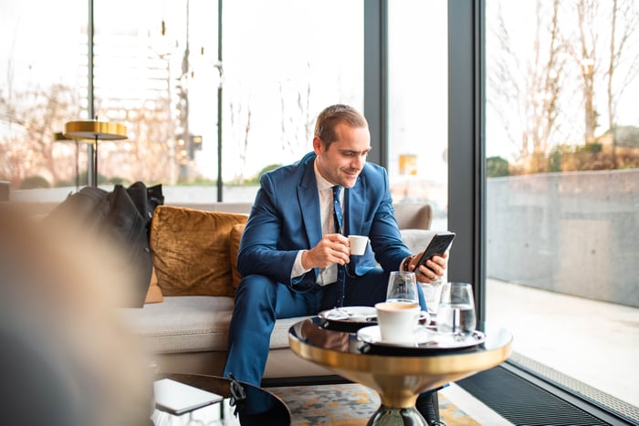 A person in suit and tie, sitting in a cafe, smiles while holding an espresso and looking at a smartphone.