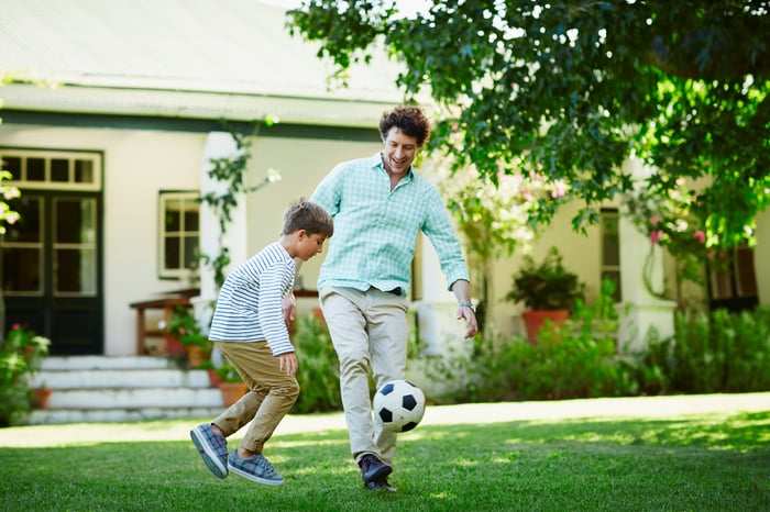 A person kicking a soccer ball with a child on the lawn in front of a house.
