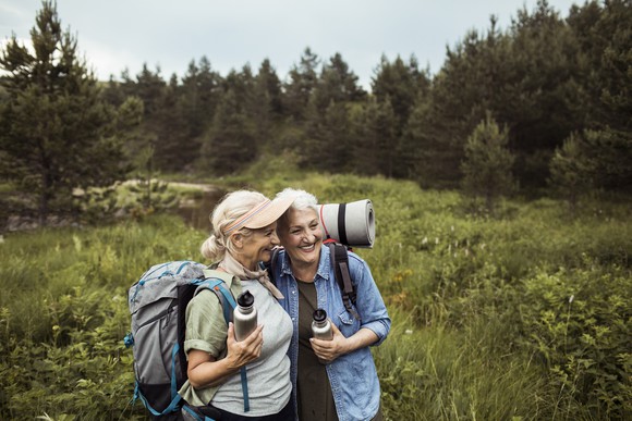 Two older adults hugging in a field while wearing hiking gear.