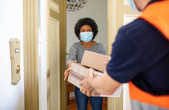 Person wearing a mask receives packages at the front door from a delivery person. 