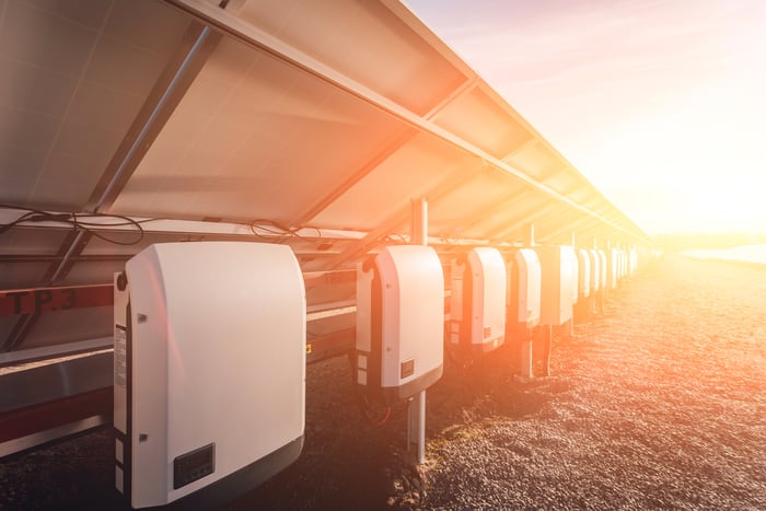 Solar inverters behind a row of solar panels in bright sunshine.