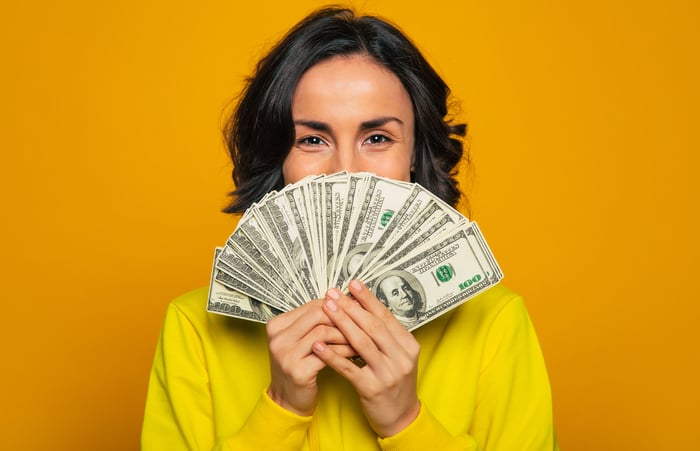 Close-up photo of a person in a yellow sweater, hiding their face behind a fan of money in their hands