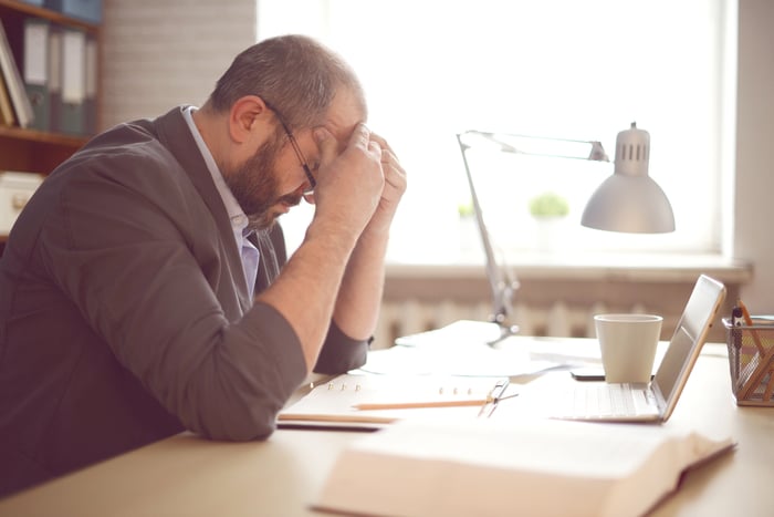 A man sits in front of a laptop and holds his head in his hands.