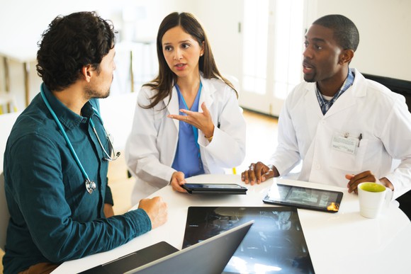 Three doctors discuss data displayed on a pair of tablets while at a desk.