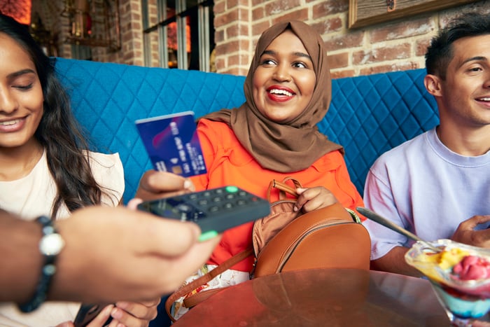 A woman wearing a hijab sitting with a man and a woman at a restaurant and paying with a credit card/