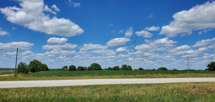 A wind power turbine in a green field, with a road in the foreground.