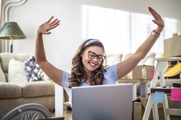 A happy person holds their arms in the air while looking at a laptop.