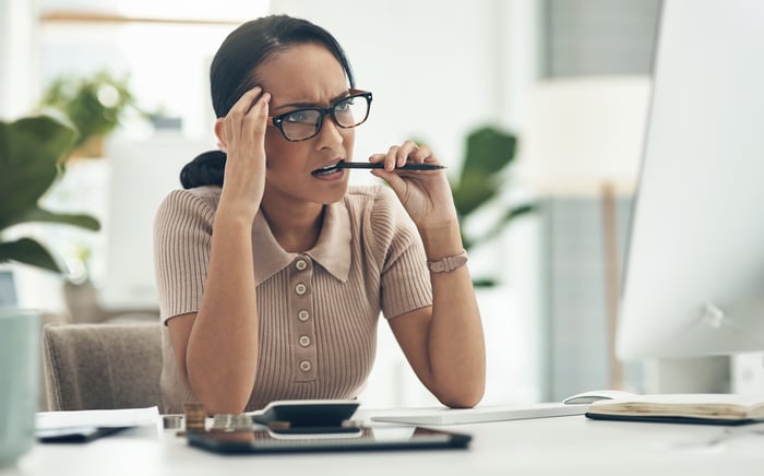 A concerned woman bites her pen and looks at a computer screen.