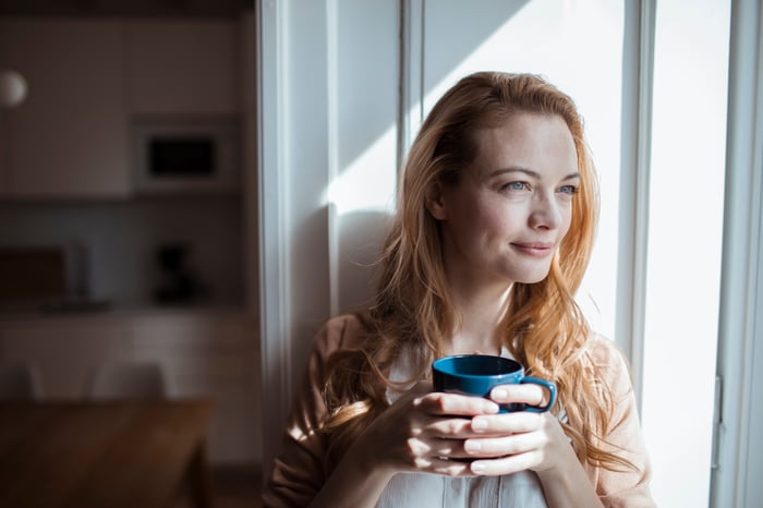 Smiling person holding coffee mug sitting in window and looking outside