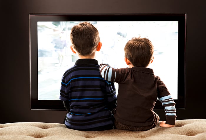 The backs of two boys sitting on a couch and watching television.