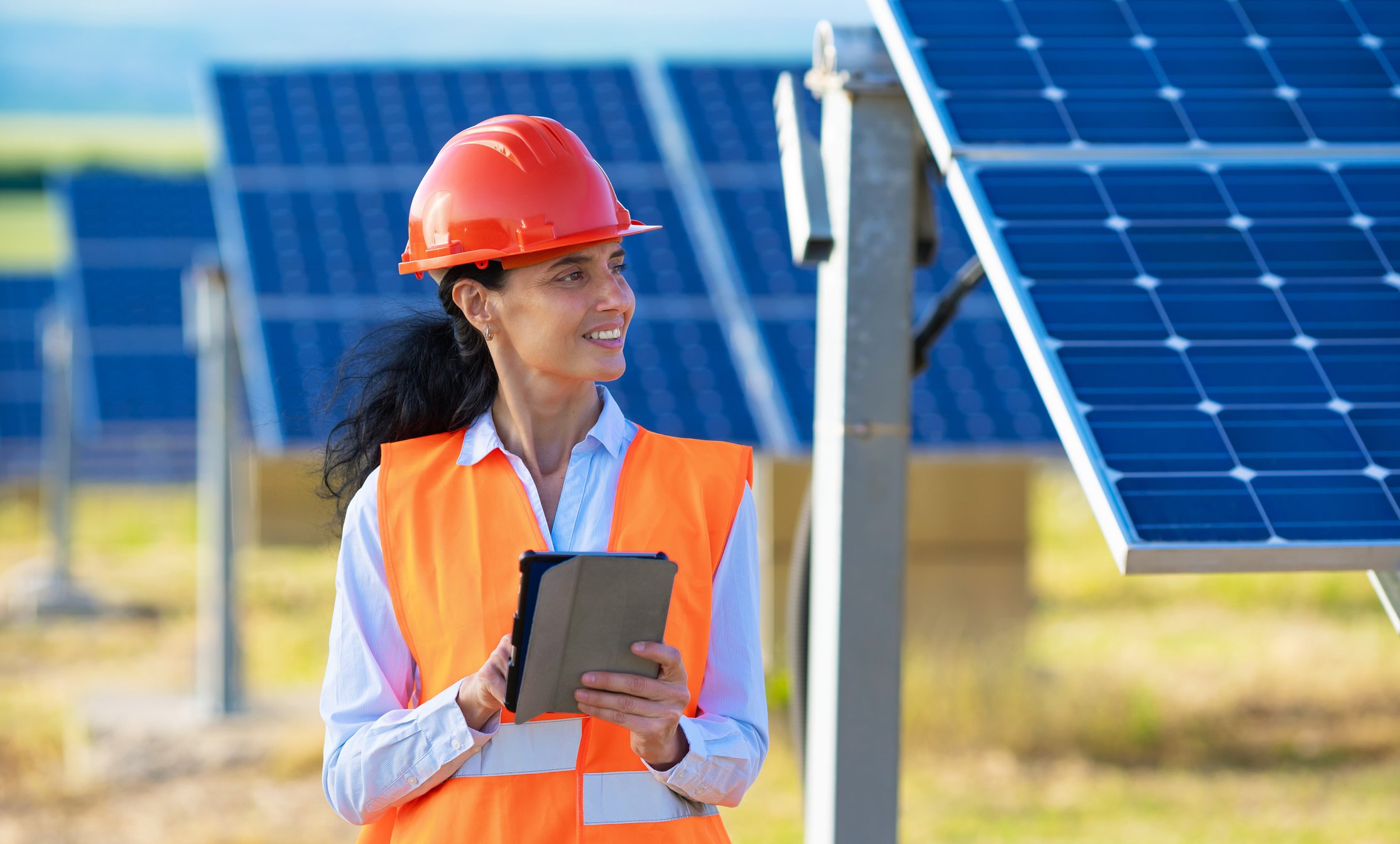 Wearing a hard hat and standing next to rows of solar panels a woman works on a tablet