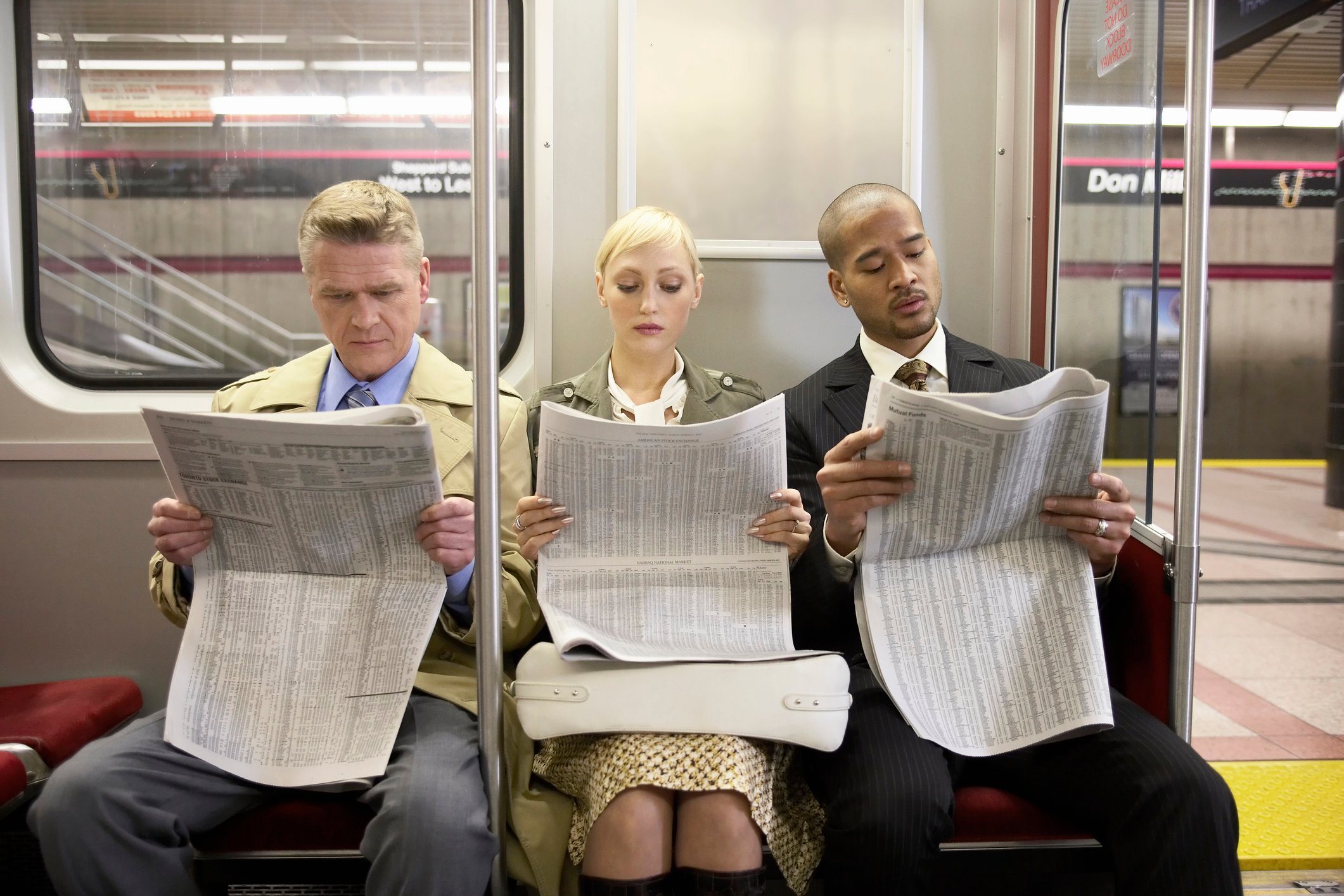 Three people sitting on a subway train reading newspaper financial pages