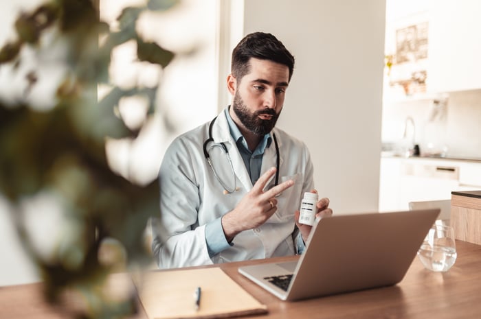 A doctor holds a bottle of pills while explaining their usage to a telemedicine patient watching them from a laptop.