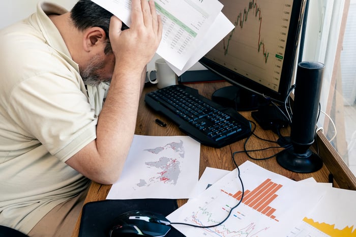 A person sits with their head in their hands at a desk with financial charts scattered on it. 