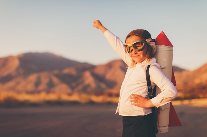 A girl with a toy rocket strapped to her back raises her arm.