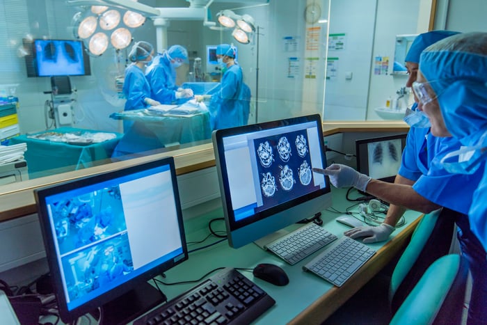 Medical technicians in a surgery control room, as doctors perform surgery in the background.