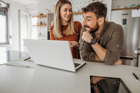 A couple sitting at the kitchen counter looking at their laptop.