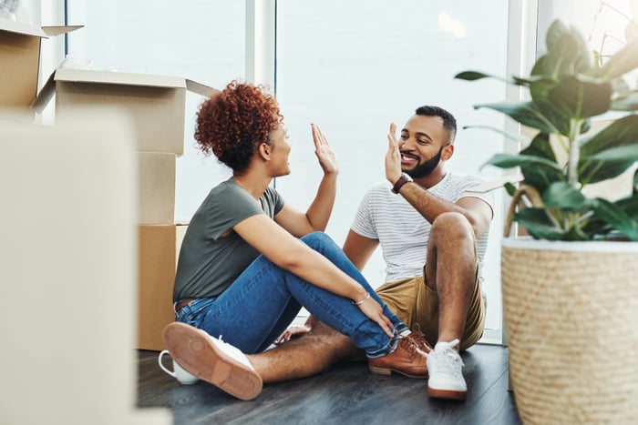 A young couple sitting on the floor of their new house and high-fiving while surrounded by boxes.