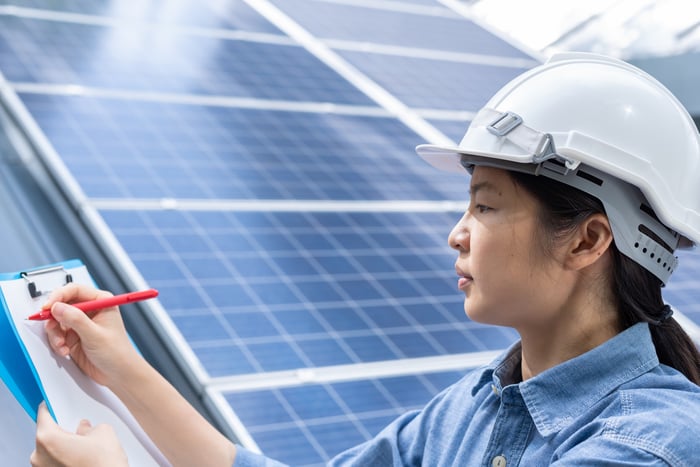 A worker writes on a clipboard while leaning against a solar panel.