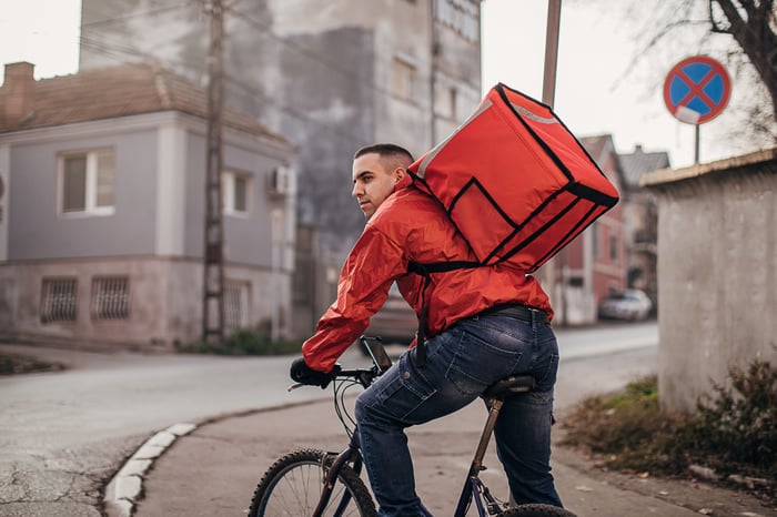 A food delivery rider with a red backpack stationary on their bike, looking around.